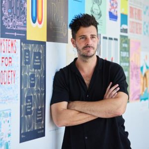 Upper body shot of Tom Cledwyn leaning, with arms crossed, against a wall covered with posters.