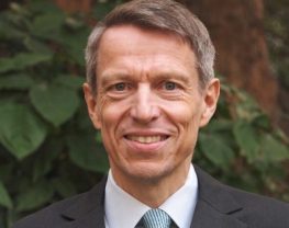 Head and shoulders shot of Soenke Ziesche smiling towards the camera, wearing a suit and tie, with a tree in the background.