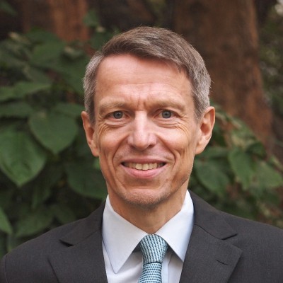 Head and shoulders shot of Soenke Ziesche smiling towards the camera, wearing a suit and tie, with a tree in the background.